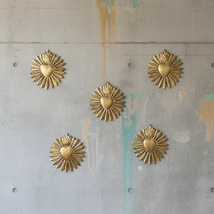 Cinq ex-voto KIFF KIFF en forme de cœur sacré en métal doré martelé disposés sur un mur en béton brut.