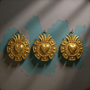 Trois ex-voto dorés KIFF KIFF modèle Ray en métal avec cœur bombé et motifs géométriques sur un mur bleu et gris