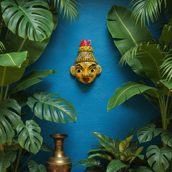 Masque mural artisanal KIFF KIFF de Krishna en papier mâché jaune, 10 cm, sur un mur bleu entouré de plantes vertes.