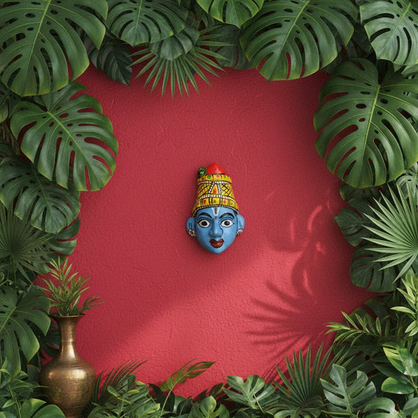 Masque mural KIFF KIFF de Radha en papier mâché bleu, peint à la main, sur un fond rouge entouré de feuilles tropicales