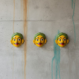 Trois masques muraux KIFF KIFF artisanaux indiens, visage jaune et coiffe verte, fixés sur un mur en béton.