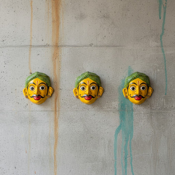 Trois masques muraux KIFF KIFF artisanaux indiens, visage jaune et coiffe verte, fixés sur un mur en béton.